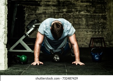 Muscular Young Man In Sweaty Shirt Training Burpee With Kettlebells On The Gym Floor With Selective Focus And Film Grain