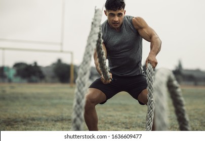 Muscular Young Man Working Out With Battling Ropes. Fit Young Male Athlete Doing Battle Rope Workout Outdoors On A Field.