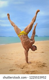 Muscular young sexy naked guy is standing on hands on beach in a speedo