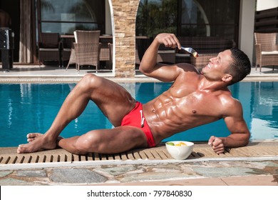 Muscular young sexy wet naked smiling guy in red speedo eating fruit near the pool outdoors