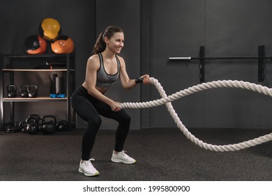 Muscular young smiling woman working out with battle ropes at dark gym, intense functional circuit training. Gray gym background with sports equipment. Crossfit, fitness and workout concept - Shutterstock ID 1995800900