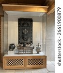 "Museum display case featuring a dark woven textile with a floral pattern, a small wooden table, and several metal pots and vessels."
