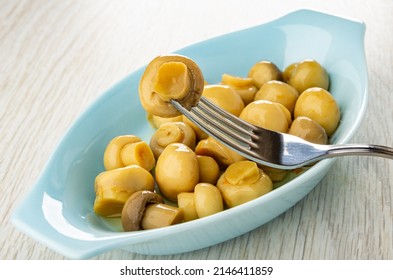 Mushroom Strung On Fork Above Blue Oval Plate With Marinated Champignons On Wooden Table