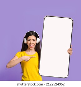 Music Application. Cheerful Asian Woman Showing Pointing Finger At Big Giant Cell Phone Blank White Screen In Hand Wearing Wireless Headphones, Violet Studio Background. Playlist App Advert Mock Up