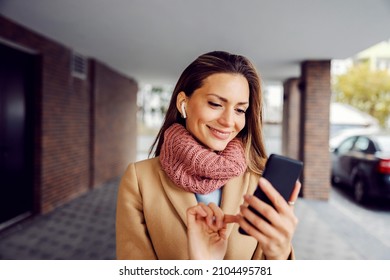 Music Apps, Technologies, And Streaming. A Happy Young Woman In A Warm Outfit Standing On The Street With Earphones And Choosing Songs On Her Phone.