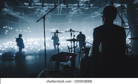 Music band group silhouette perform on a concert stage.  
				silhouette of drummer playing on drums
				audience holding cigarette lighters and mobile phones