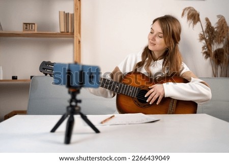 Music college student practicing acoustic guitar exercise, recording video on phone on tripod. Woman taking an online musical courses at home. Copy space