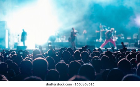 Music Fans Recording Videos On Phone In Crowd On Concert, Rear Back View Of Audience People Using Devices Enjoy Live Music Festival Event Shooting Rock Band Stage On Mobile Device In Blue Lights.