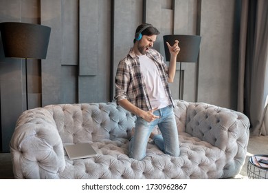 Music, Life. Enthusiastic Man In Headphones Playing An Air Guitar On His Knees On The Couch, Singing Happy.