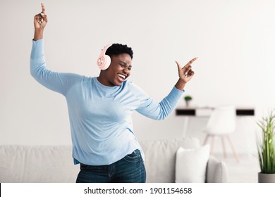 Music Lover Concept. Overjoyed ecstatic plus-size african american lady wearing pink wireless headphones listening to favourite song, dancing at home with closed eyes. Female taking break, copy space - Powered by Shutterstock