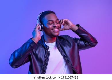Music Lover. Handsome Black Guy In Wireless Headphones And Leather Jacket Listening His Favorite Songs, Relaxed African American Man Standing With Closed Eyes Under Neon Light Over Purple Background - Shutterstock ID 1931037413