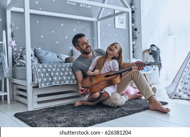                                Music is so much fun. Young father teaching his little daughter to play guitar and smiling while sitting on the floor in bedroom - Powered by Shutterstock