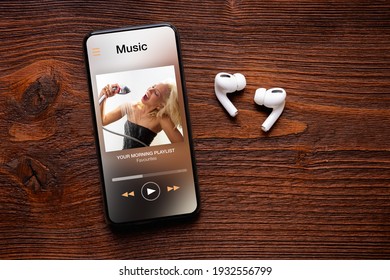 Music player on screen of mobile phone and wireless earbuds on wooden surface