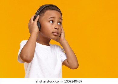 Music, Relaxation, Fun, Joy And Modern Electronic Gadgets Concept. Adorable Dark Skinned Black Male Kid Enjoying Favorite Tracks Or Listening To Radio Via Wireless Headphones, Looking Upwards