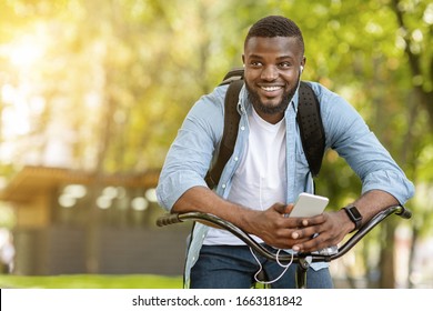 Music For Ride. Portrait Of Happy Afro Man Sitting On Bike And Listening His Favorite Songs On Smartphone With Earphones, Free Space