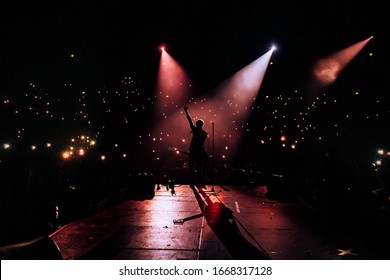 Music Show. Guitarist In Front Of Crowd On Scene In Night Club. Bright Stage Lighting, Crowded Dance Floor. Phone Lights At Concert. Band Red Silhouette Crowd. People With Cell Phone 