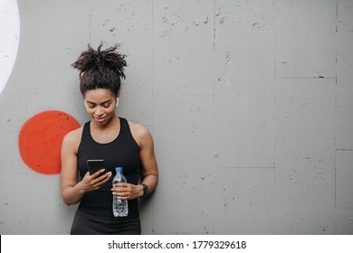 Music And Sport. Smiling Muscular African American Young Woman With Headphones, Smart Watch And Bottle Of Water Looks At Smartphone On Wall Background, Free Space