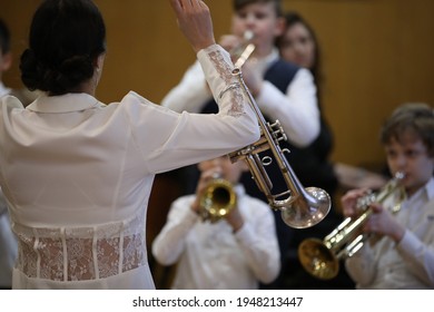 A music teacher a woman in a white dress an orchestra conductor with her hands raised stands in front of young musicians playing a musical instrument trumpet a concert at a children's school.Creation