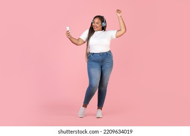 Musical App. Black Plus Size Woman Listening To Music In Headphones Holding Phone Dancing And Having Fun Enjoying Favorite Song Posing Over Pink Studio Background. Full Length Shot - Powered by Shutterstock