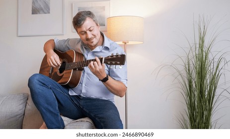 Musical hobby. Guitar skill. Happy smiling man sitting on sofa playing string instrument enjoying learning chords in living room interior at home copy space. - Powered by Shutterstock