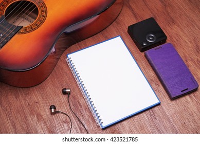 Musical Theme. Blank White Spiral Bound Book, Earphone, Partial View Of A Guitar, A Square Shaped Media Player And A Purple Colored Media Player. 