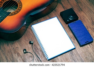 Musical Theme. Blank White Spiral Bound Book, Earphone, Partial View Of A Guitar, A Square Shaped Media Player And A Purple Colored Media Player. Rustic Tone Image Processing Technique Applied. 