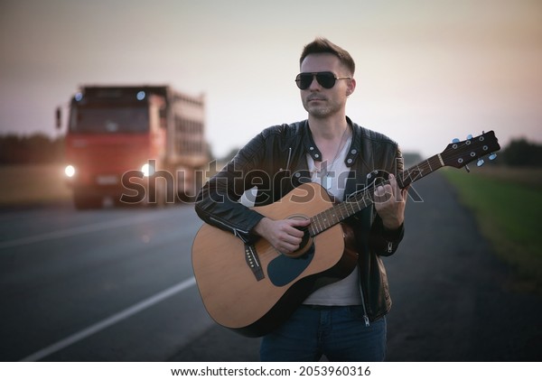 A musician\
with the guitar stands on the\
road.