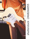 A musician in an orange plaid shirt plays a white electric guitar, possibly performing at an outdoor concert