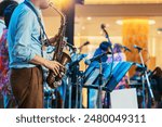 Musician playing saxophone on blurred background. Man with friends blow saxophone with the band for performance. Music instrument played by saxophonist player musician in festival. Selective focus.