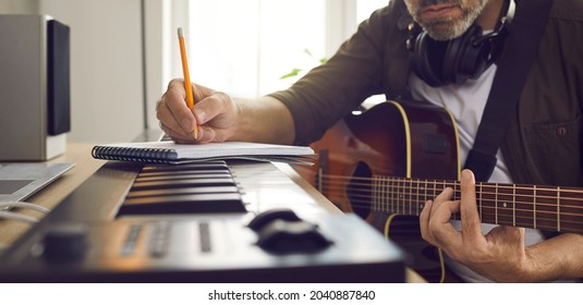 Musician writing music. Songwriter who's following his passion sitting by electronic keyboard in home studio, holding pencil, picking guitar strings, thinking of lyrics for new song. Banner background - Shutterstock ID 2040887840