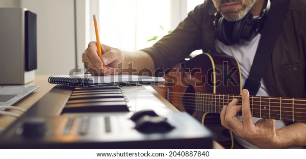 Musician writing music. Songwriter who\'s\
following his passion sitting by electronic keyboard in home\
studio, holding pencil, picking guitar strings, thinking of lyrics\
for new song. Banner\
background