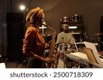 Musicians rehearsing jazz performance with saxophonist in foreground and drummer in background. Recording studio setting with instruments and equipment surrounding musicians