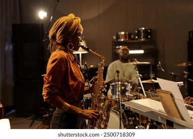 Musicians rehearsing jazz performance with saxophonist in foreground and drummer in background. Recording studio setting with instruments and equipment surrounding musicians - Powered by Shutterstock