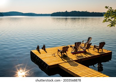 Muskoka or Adirondack chairs sitting on a wooden dock facing lake at sunset - sunrise, long wooden pier. Surrounded by green trees and hills