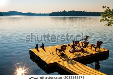 Muskoka or Adirondack chairs sitting on a wooden dock facing lake at sunset - sunrise, long wooden pier. Surrounded by green trees and hills