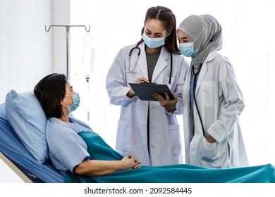 Muslim Arab Islam And Asian Intern Doctors In White Lab Coat With Hijab Face Mask And Stethoscope Discussing Talking Consulting Infected Coronavirus Patient Checklist Symptom On Clipboard Together.