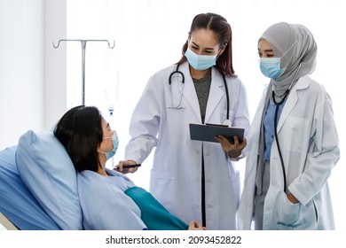 Muslim Arab Islam And Asian Intern Doctors In White Lab Coat With Hijab Face Mask And Stethoscope Discussing Talking Consulting Infected Coronavirus Patient Checklist Symptom On Clipboard Together.