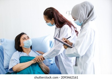 Muslim Arab Islam And Asian Intern Doctors In White Lab Coat With Hijab Face Mask And Stethoscope Discussing Talking Consulting Infected Coronavirus Patient Checklist Symptom On Clipboard Together.