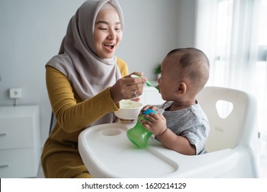 Muslim Asian Mother Feeding Her Baby Boy While Sitting On High Chair At Home