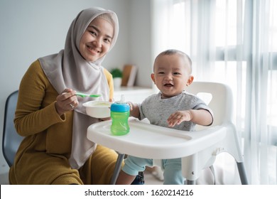 Muslim Asian Mother Feeding Her Baby Boy While Sitting On High Chair At Home