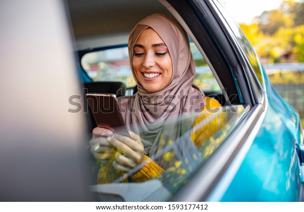 Muslim business\
woman in car. Writing a message on her mobile phone. Positive\
pensive Islamic woman in hijab sitting on backseat of taxi and\
drinking coffee while using\
gadget