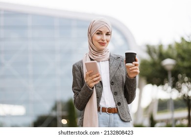 Muslim business woman in hijab standing in front of modern building or airport, smiling and talking on cell phone, drinking take away coffee - Powered by Shutterstock