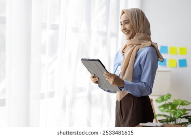 Muslim businesswoman holding tablet and smiling in office. Young businesswoman is holding a tablet and smiling in a modern office. She is wearing a hijab and professional attire. - Powered by Shutterstock