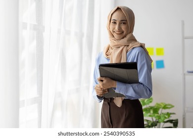 Muslim businesswoman holding tablet and smiling in office. Young businesswoman is holding a tablet and smiling in a modern office. She is wearing a hijab and professional attire. - Powered by Shutterstock