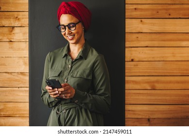 Muslim Businesswoman Using A Her Smartphone While Standing Against A Wall. Cheerful Businesswoman Smiling While Sending A Text Message. Female Entrepreneur Wearing A Headscarf In A Modern Workplace.