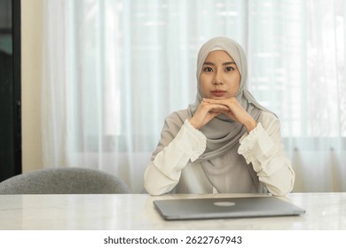 Muslim confidence woman in hijab, leadership, serious asian muslim businesswoman wearing light hijab sits at modern office desk with laptop, looking straight, calm focused mindset at home