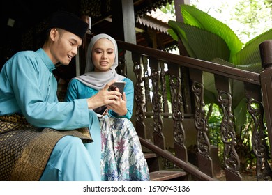  Muslim Couple In Traditional Malay Clothing Sitting At Wooden Stair Using Smart Phone During Eid Al-Fitr Celebration.