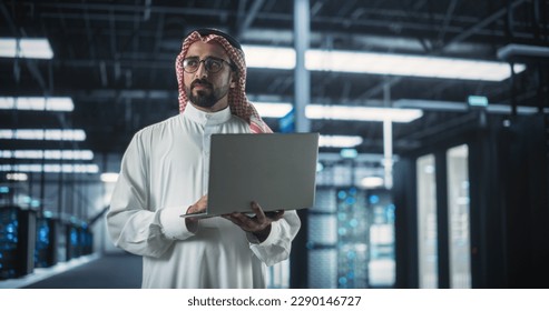 Muslim Data Center IT Engineer Standing in a Room with Operational Server Racks. Cloud Computing Architect Uses Laptop Computer for Servicing the System in Cyber Security and Data Protection Facility