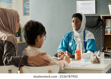 Muslim doctor discussing treatment with patient and family member in modern medical office. Prescription bottles and files on desk, creating supportive environment - Powered by Shutterstock