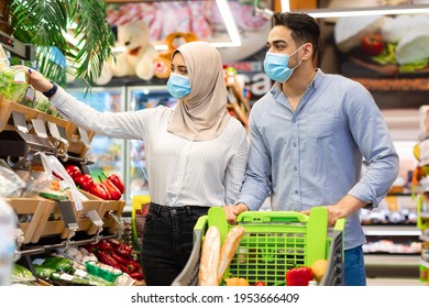 Muslim Family Couple Doing Grocery Shopping Wearing Protective Face Masks Buying Food Together In Modern Supermarket. Islamic Husband And Wife Choosing Fresh Organic Vegetables In Hypermarket